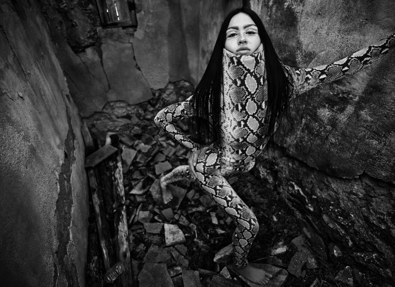 Woman in a snakeskin-patterned suit strikes a pose in front of a crumbling wall. Black and white portrait in an industrial setting.