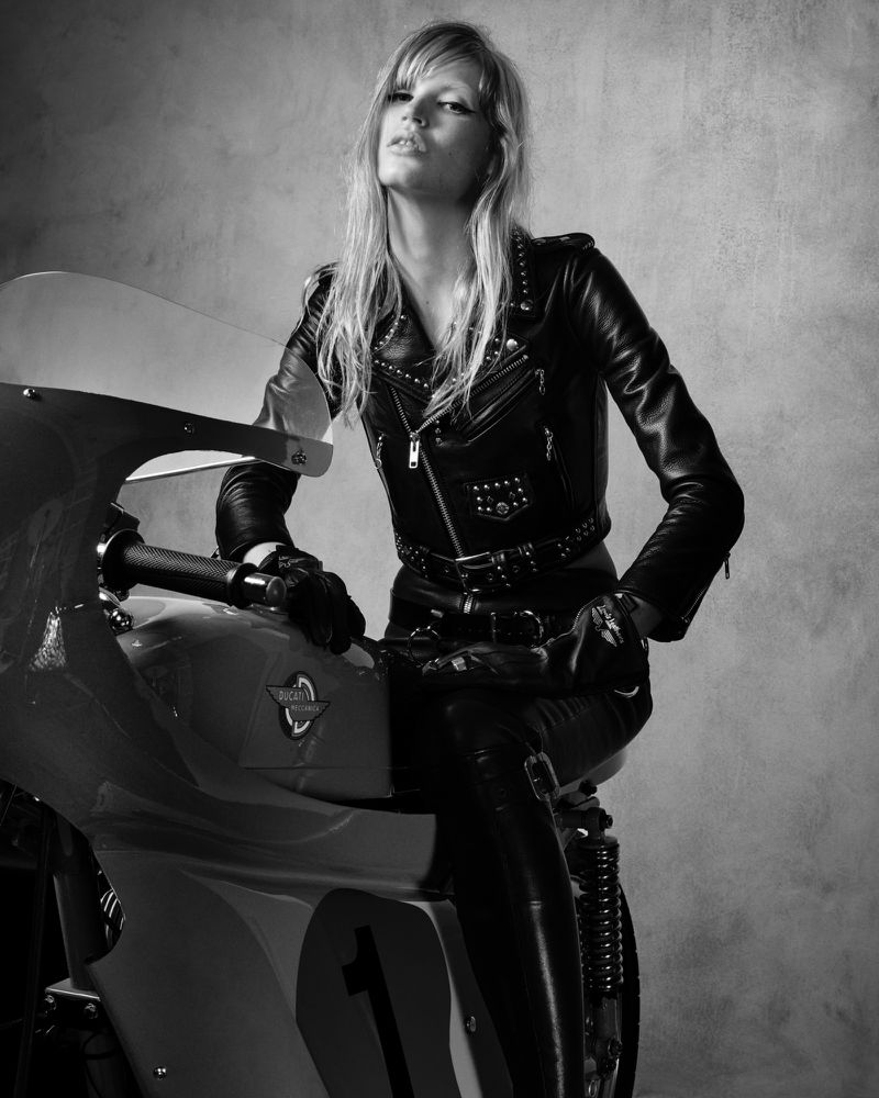 Monochrome shot of a blonde adult female in black leather jacket and leggings sitting on a vintage motorcycle against a textured background.