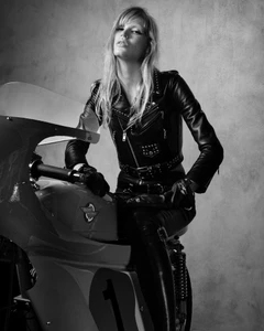 Model Jacqui Hooper posing on a vintage motorcycle wearing a black leather jacket against a textured grey background.