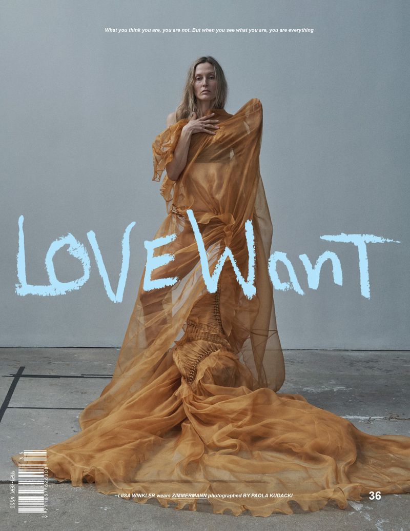 A woman poses wearing a sheer amber couture gown in front of a pale grey backdrop and a handwritten-font logo in a fashion editorial.