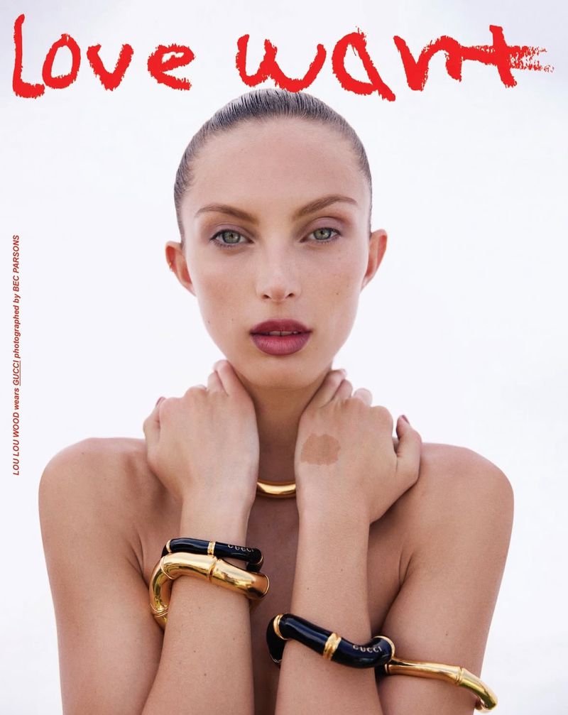 A model poses with gold tone jewelry against a white background with red painted text reading LOVE WANT in a high fashion setting.
