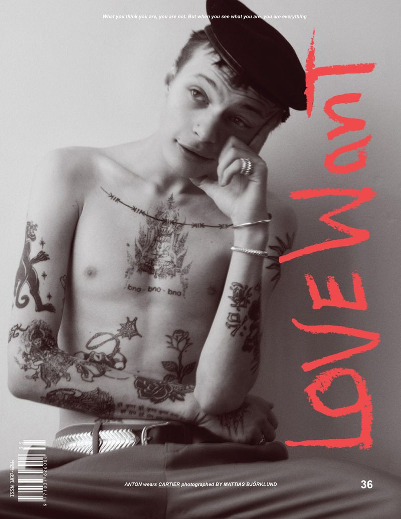 Tattooed man in a beret poses topless indoors, seated against a neutral wall with bright lighting. Red text overlay.