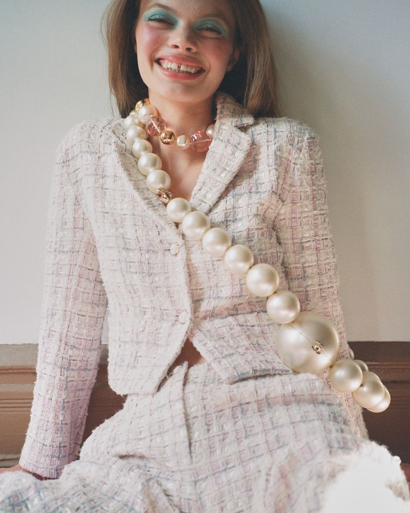 A young woman with bright makeup smiles while wearing a tweed suit and a chunky beaded necklace in a bright, indoor setting.