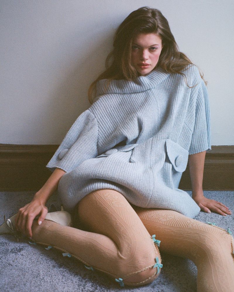A young woman poses indoors wearing a light-blue knitted sweater dress and textured tights with bows, set against a neutral wall and floor.