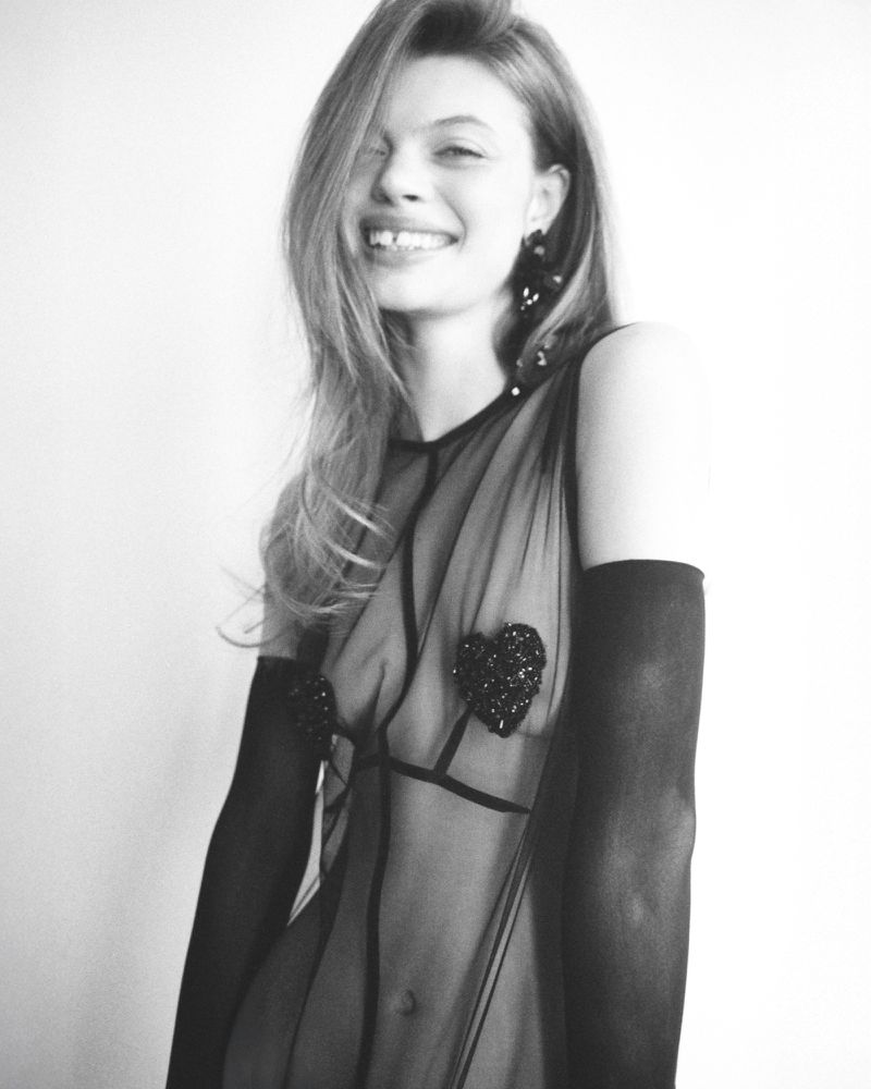 A smiling woman with long hair wearing a black see-through dress with heart appliques, black gloves, and large earrings stands against a white wall.