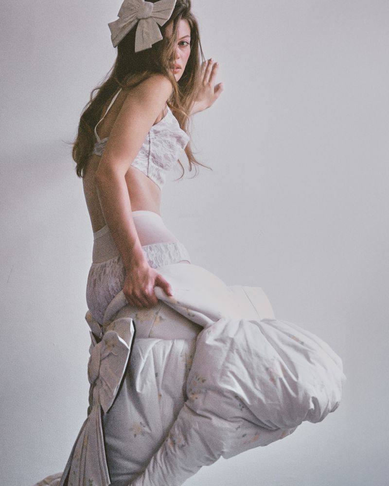 Woman in lingerie poses with duvet, wearing a large bow in her hair against a plain white background in a studio-like setting.