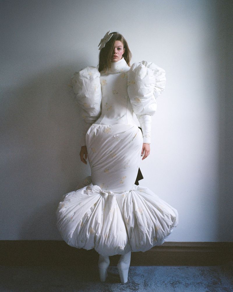 Fashion portrait of a young woman in a puffy white dress, white tights, and sculptural white hair accessory in a minimalist studio.