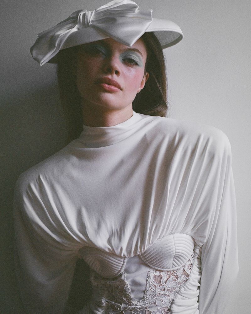 Fashion shot featuring a female model in a white dress and large bow hat. Soft lighting and pale colours create a delicate aesthetic.