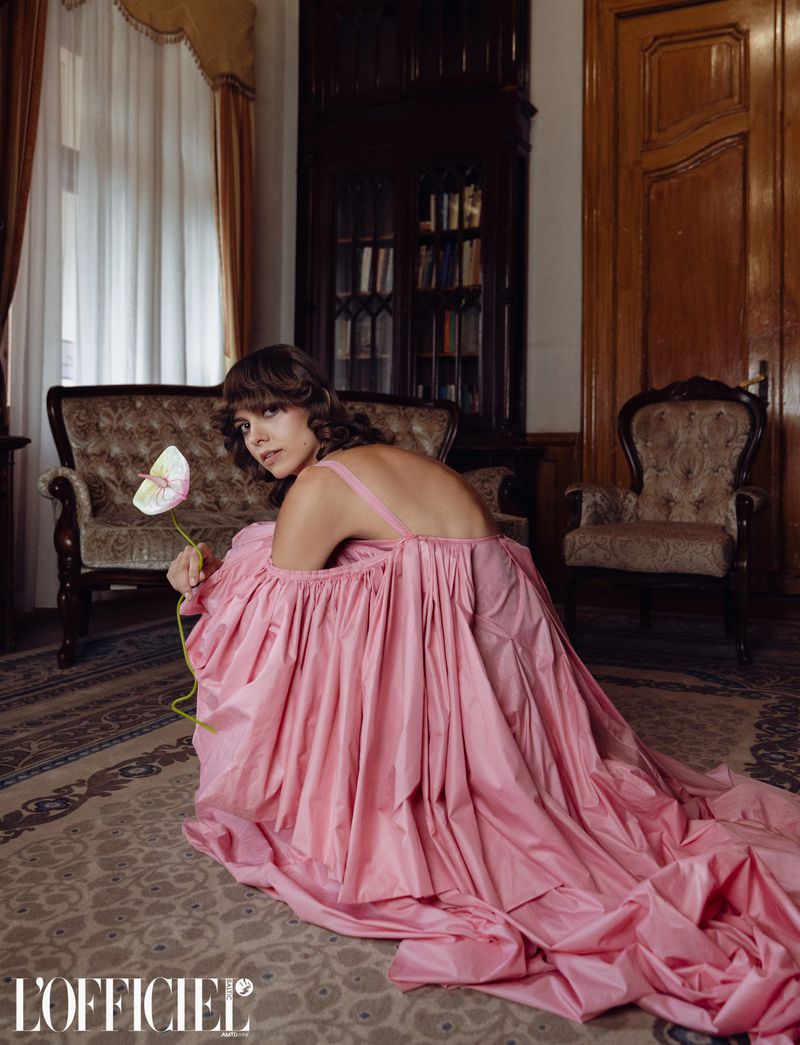 A young woman in a pink gown squats holding a flower, looking at the camera indoors, with vintage furniture and a floral rug.