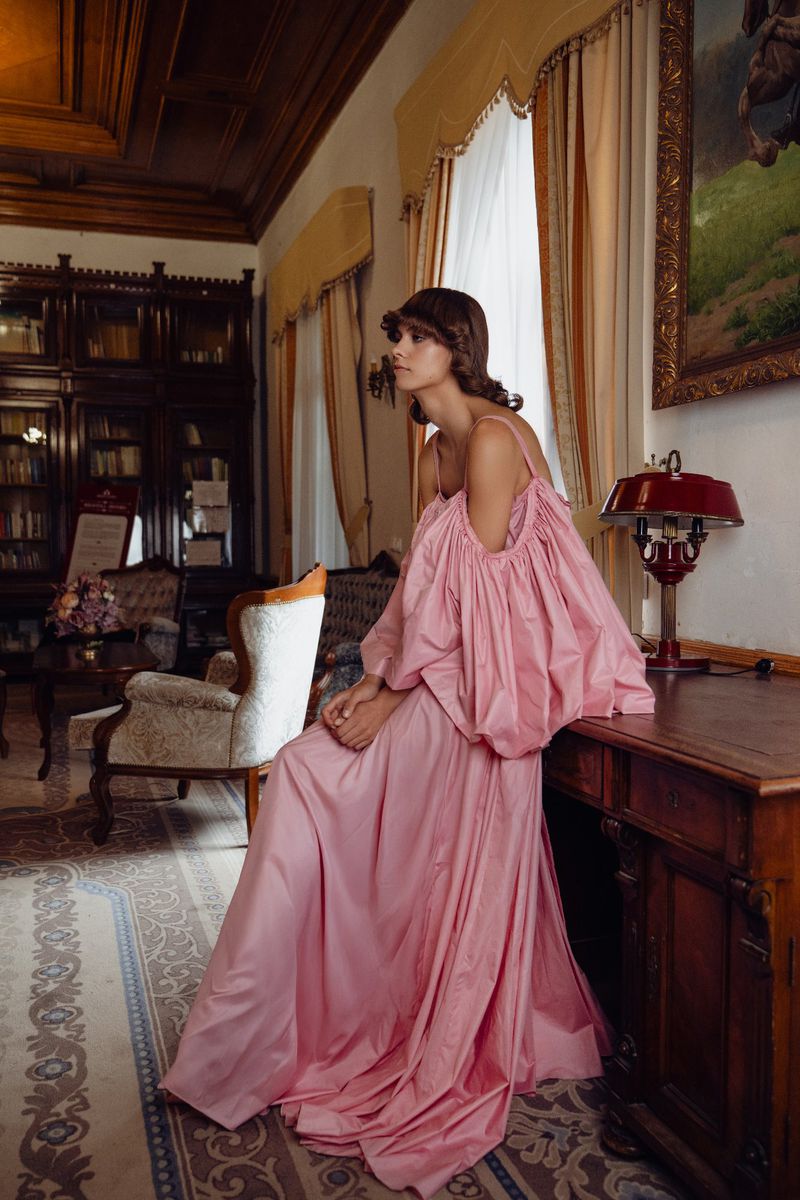 Woman in a flowing pink dress poses in a room with a bookcase and ornate furniture, bathed in soft, natural light.