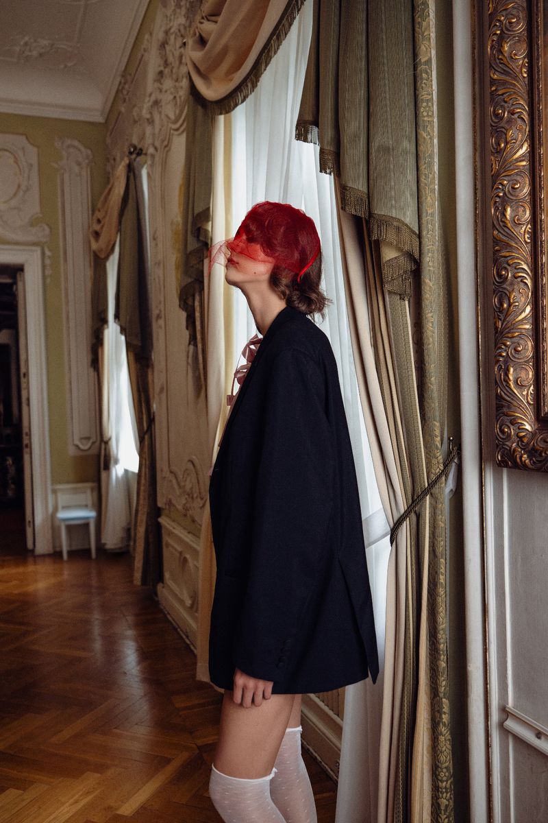 Woman in oversized blazer and stockings poses by window in ornate room with draped curtains and gilded wall decor; filtered indoor light.