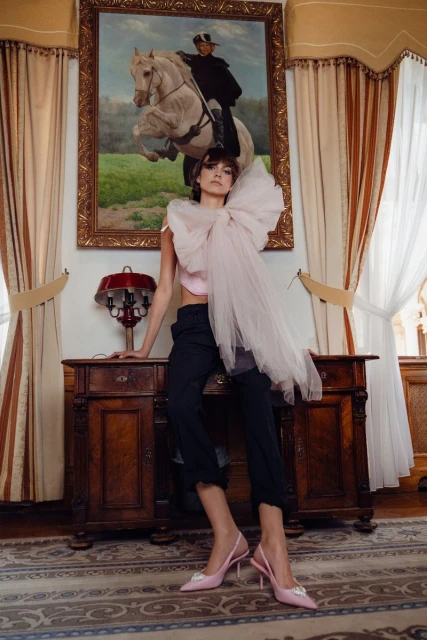 Fashionable woman in pink top and shoes poses near wooden desk and painting of horse rider in luxurious indoor setting.