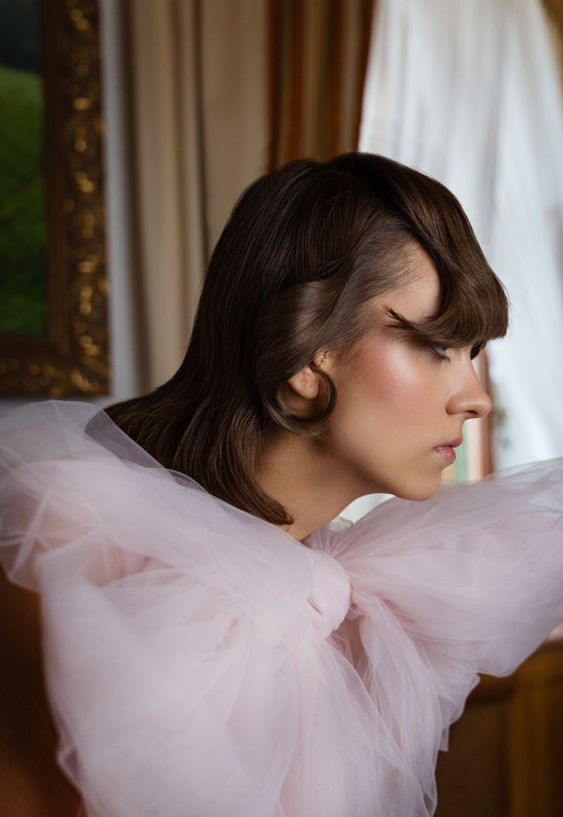 Profile of a woman with brown wavy hair, wearing pink tulle garment, posing indoors with natural light, in front of a framed painting.