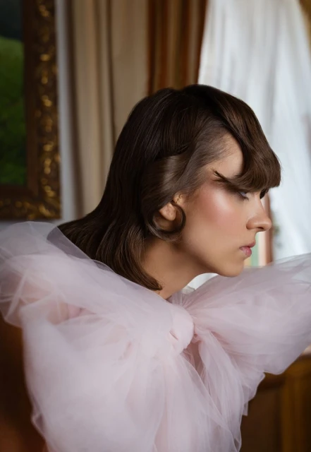 Profile of a woman with brown wavy hair, wearing pink tulle garment, posing indoors with natural light, in front of a framed painting.