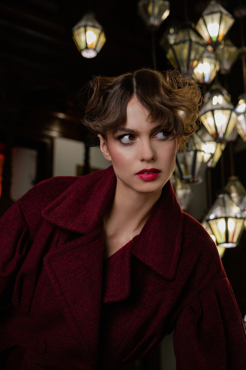 Portrait of a young woman with red lipstick wearing a maroon coat, posing in an indoor setting with a backdrop of decorative lights.