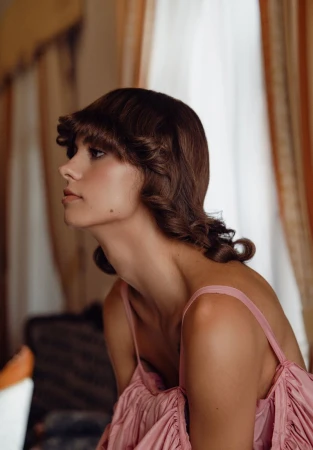 Woman with brown hair in pink dress with curtain background looks left in a beauty shot showcasing hair and clothing.