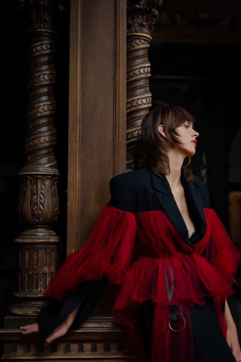 A woman poses in a black blazer with red tulle sleeves next to an ornate wooden column, side lit in a dark interior.