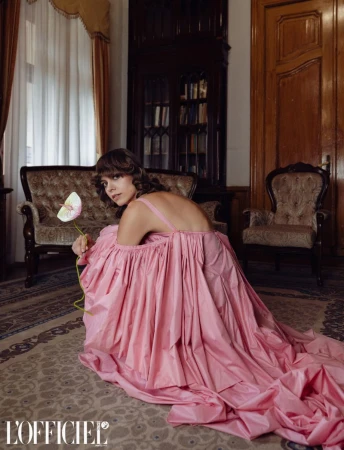 A woman in a voluminous pink dress holding a flower, kneeling on a patterned rug inside an ornate room with vintage furniture.