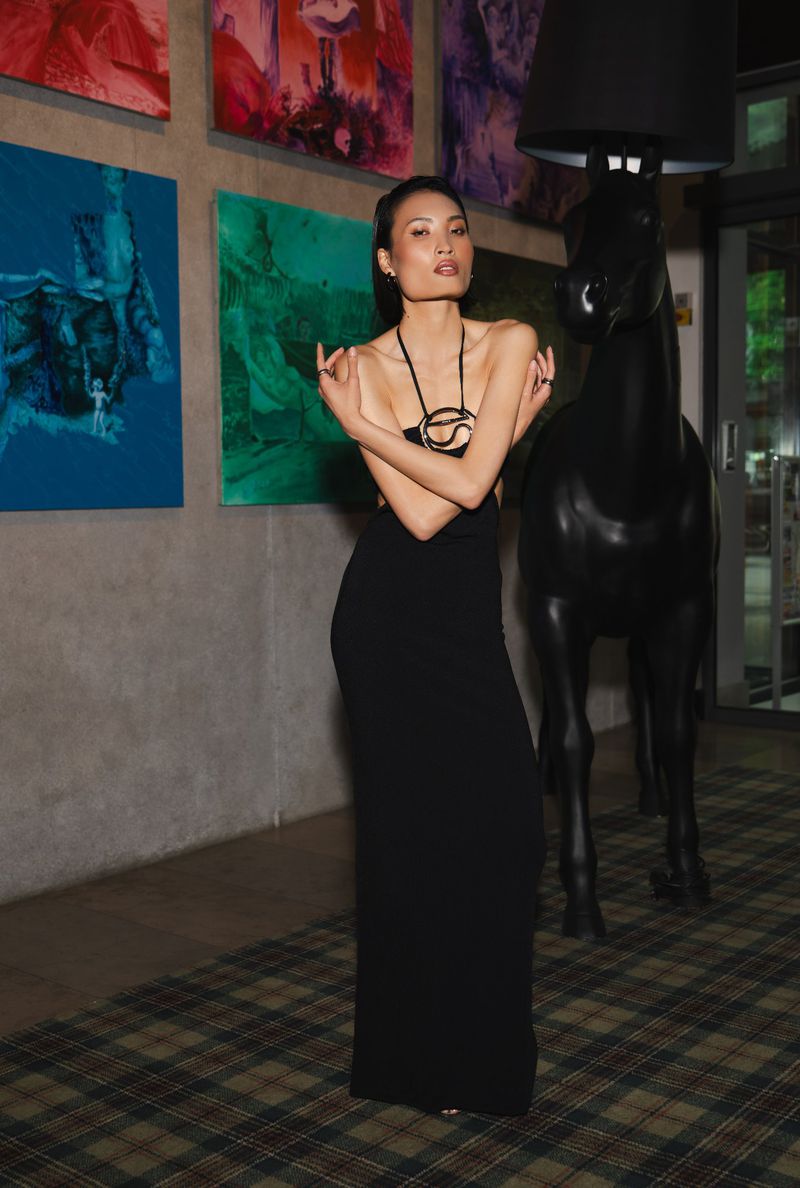 Elegant Asian woman in a black strapless dress and statement necklace, posing next to a black horse sculpture in an indoor setting.