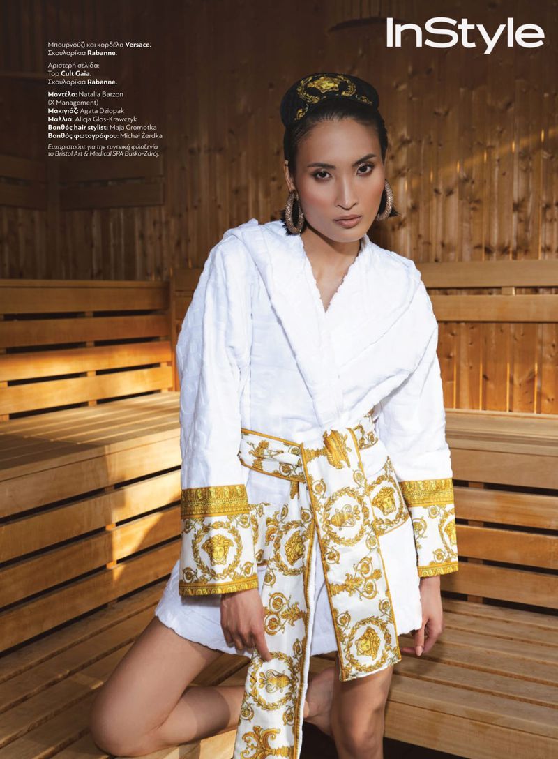 Editorial fashion shot of a woman in a white robe and headscarf with a gold Versace pattern in a sauna with wood benches.