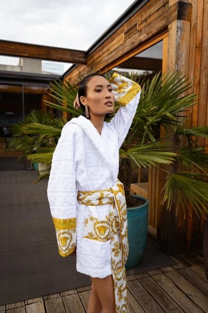 Woman in luxurious white Versace bathrobe standing outdoors on wooden deck with tropical plants in background, cloudy sky overhead.