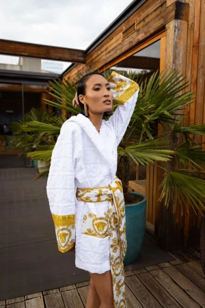 Woman in luxurious white Versace bathrobe standing outdoors on wooden deck with tropical plants in background, cloudy sky overhead.