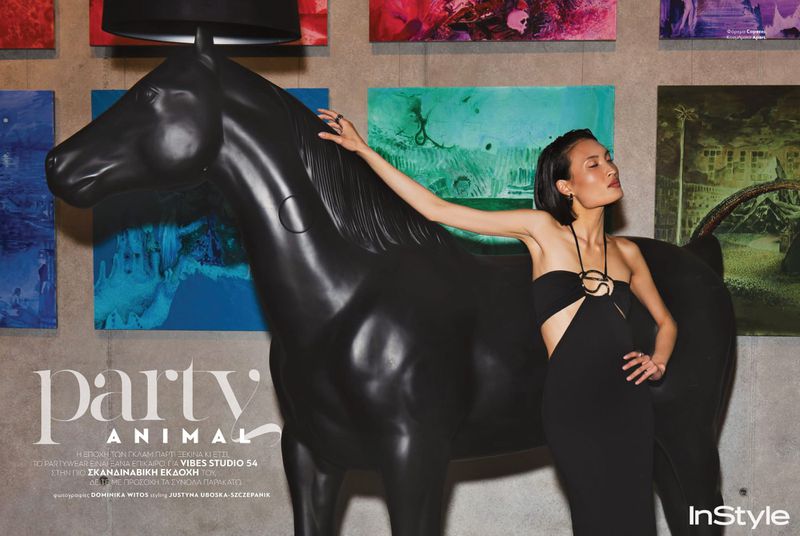 A woman in a black dress poses next to a black horse statue with a lamp, in front of a wall displaying colorful, diverse painting art pieces.