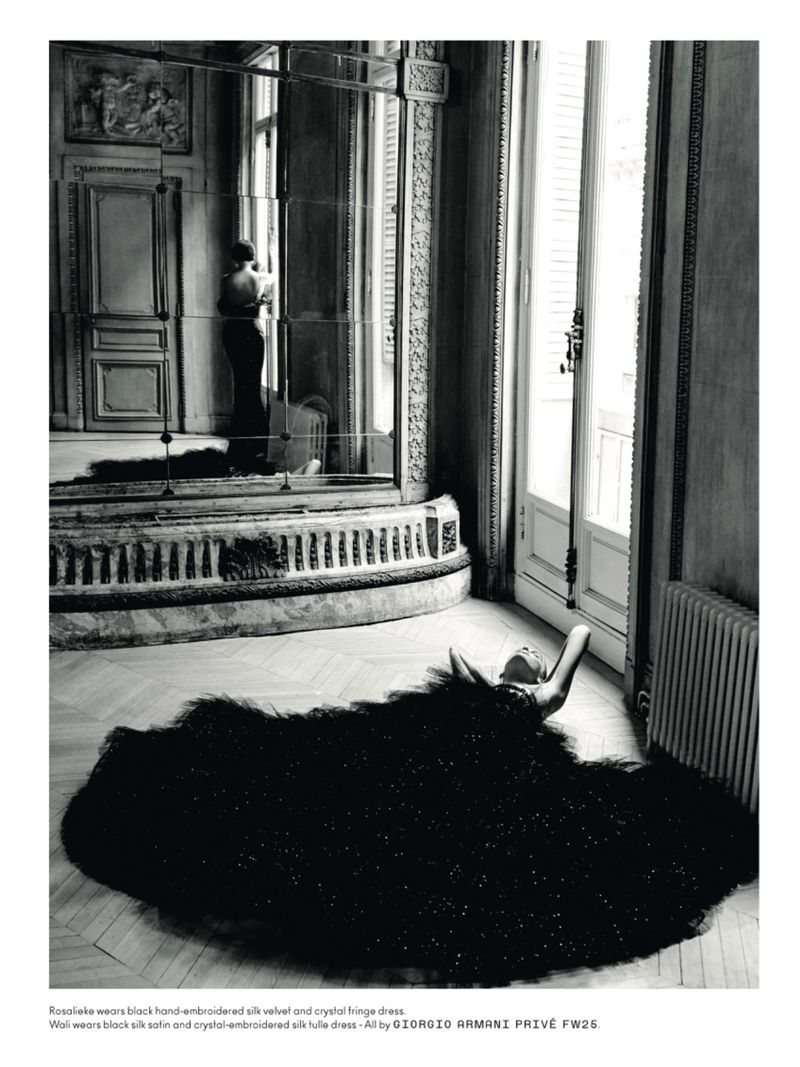 Fashion editorial shot in black and white: a woman lies in a black gown, while another woman looks out the window in a grand room.