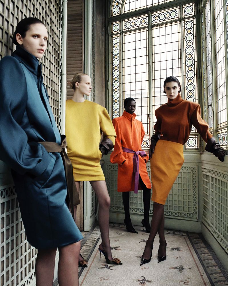 Four fashion models in bright color-blocked dresses and coats pose inside a light-filled vintage building with tiled floors.