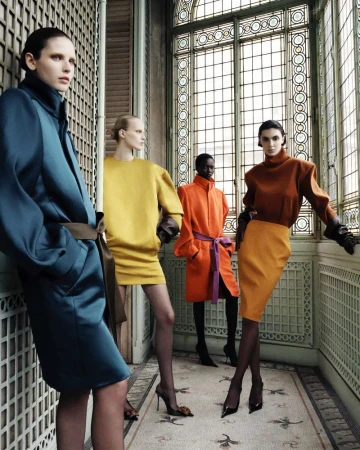 Four fashion models in bright color-blocked dresses and coats pose inside a light-filled vintage building with tiled floors.