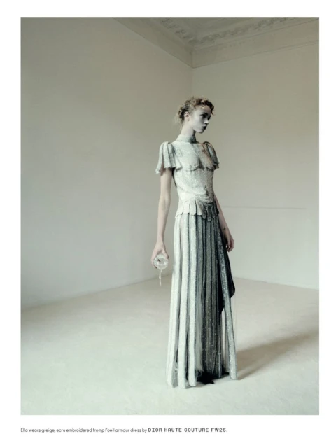Fashion shot of a female model wearing a long, formal dress with lace details in a minimalist photo studio with high ceilings.