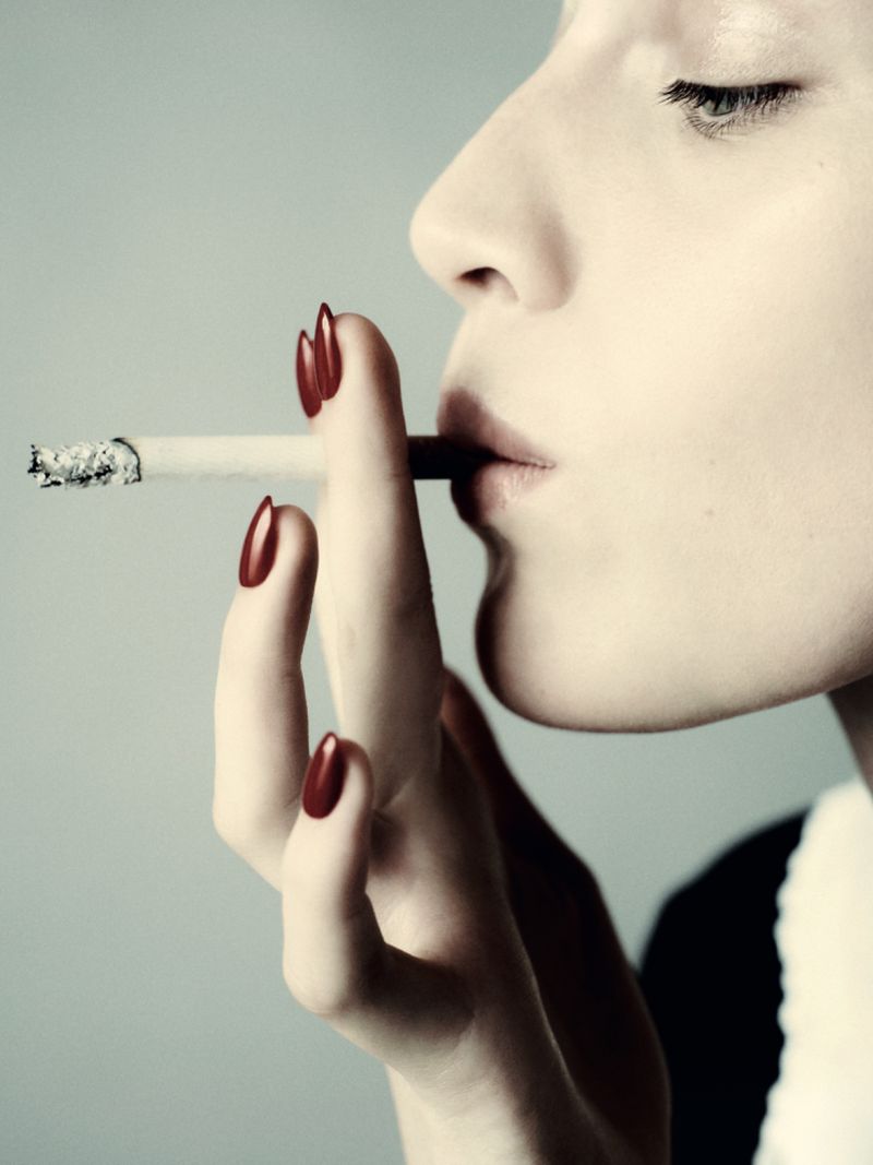 Woman with red nails and light skin tone smoking a cigarette in a close-up shot with a neutral gray background.