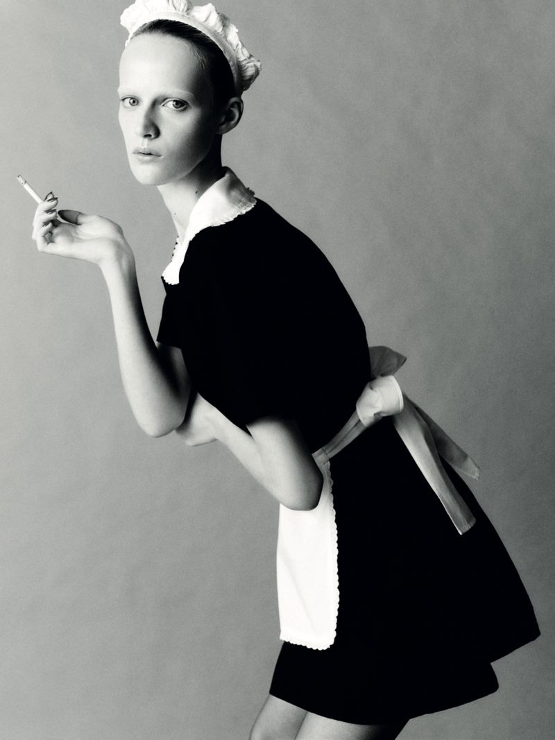 Black and white portrait of a model in a maid outfit holding a cigarette, set against a simple grey backdrop in a studio.