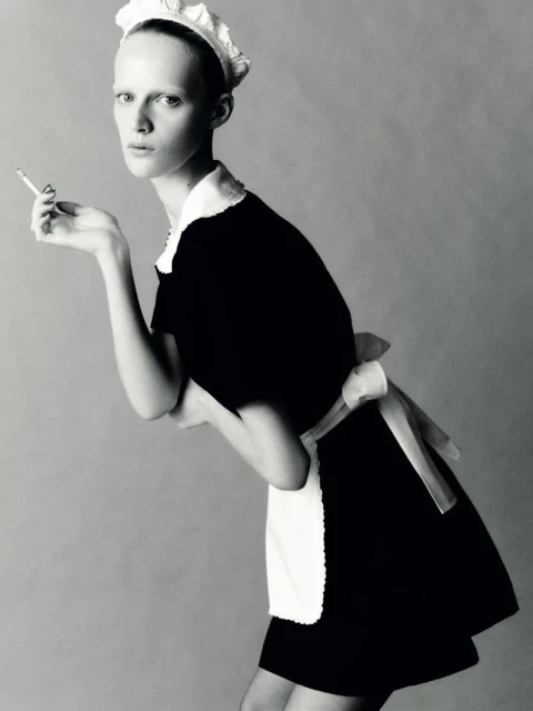 Black and white portrait of a model in a maid outfit holding a cigarette, set against a simple grey backdrop in a studio.