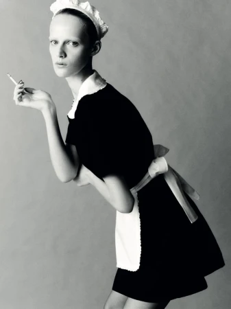Black and white portrait of a model in a maid outfit holding a cigarette, set against a simple grey backdrop in a studio.