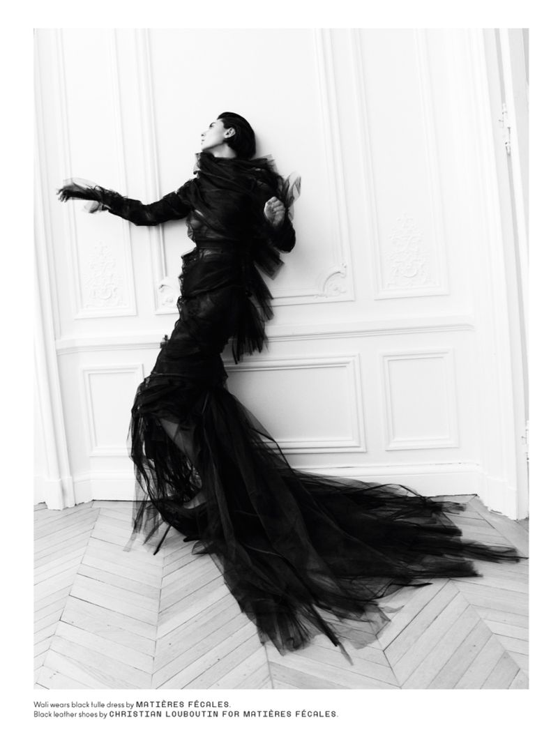 Woman in black tulle dress poses dramatically against a white paneled wall, showcasing fashion design in a high-contrast black and white image.