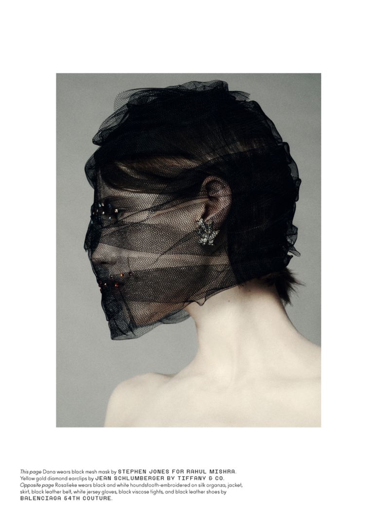 Woman in a black mesh mask and jewelry, shot in a studio with neutral grey backdrop. Fashion editorial style.