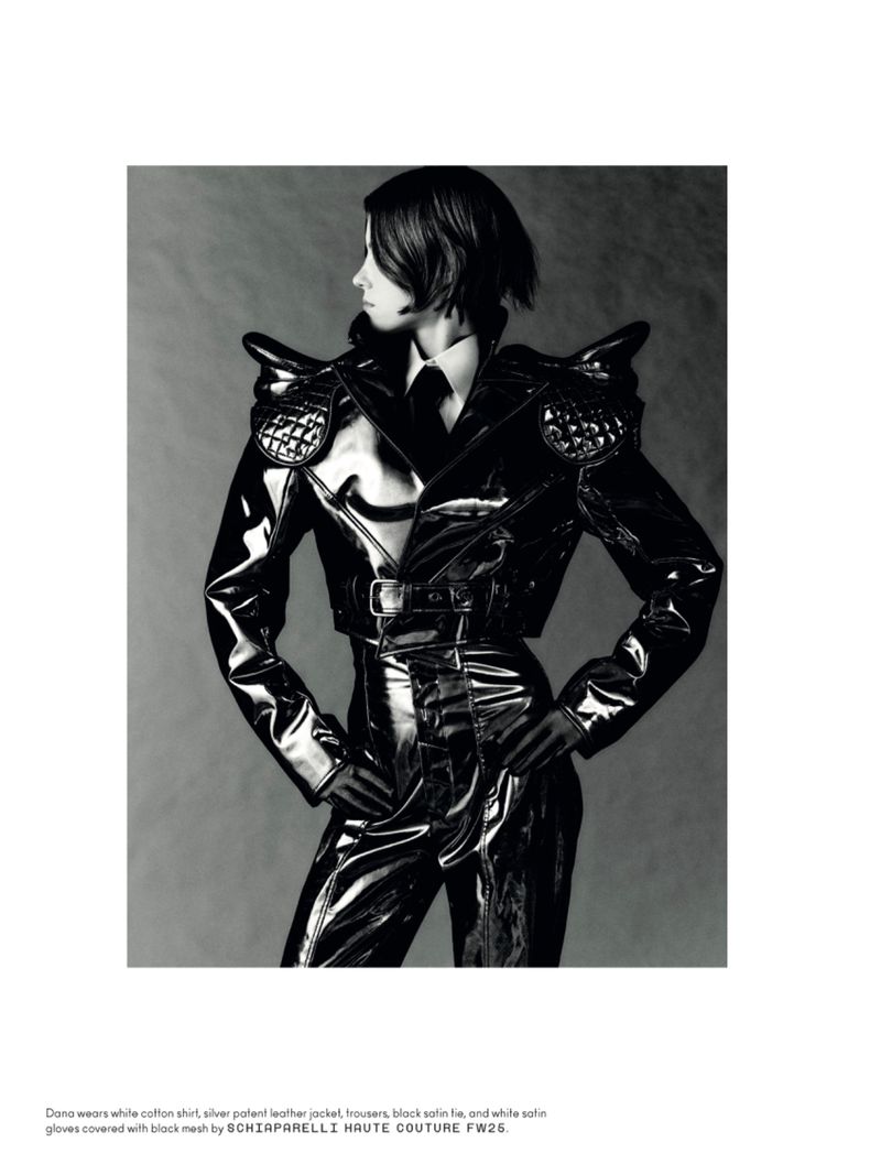 A woman poses in black shiny clothing with padded shoulders against a gray background, creating a high-fashion editorial look.