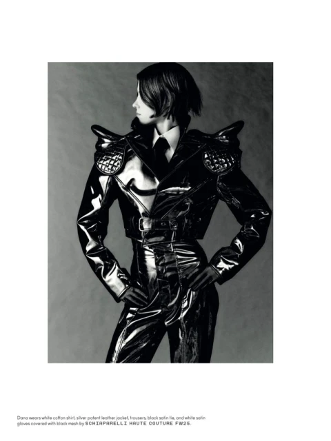 A woman poses in black shiny clothing with padded shoulders against a gray background, creating a high-fashion editorial look.
