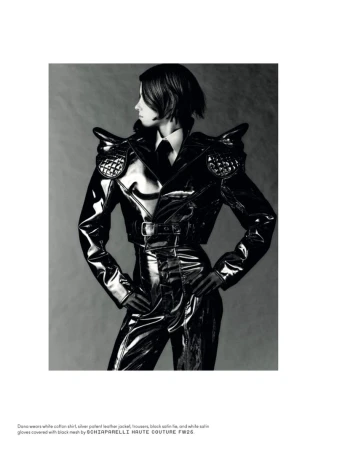 A woman poses in black shiny clothing with padded shoulders against a gray background, creating a high-fashion editorial look.