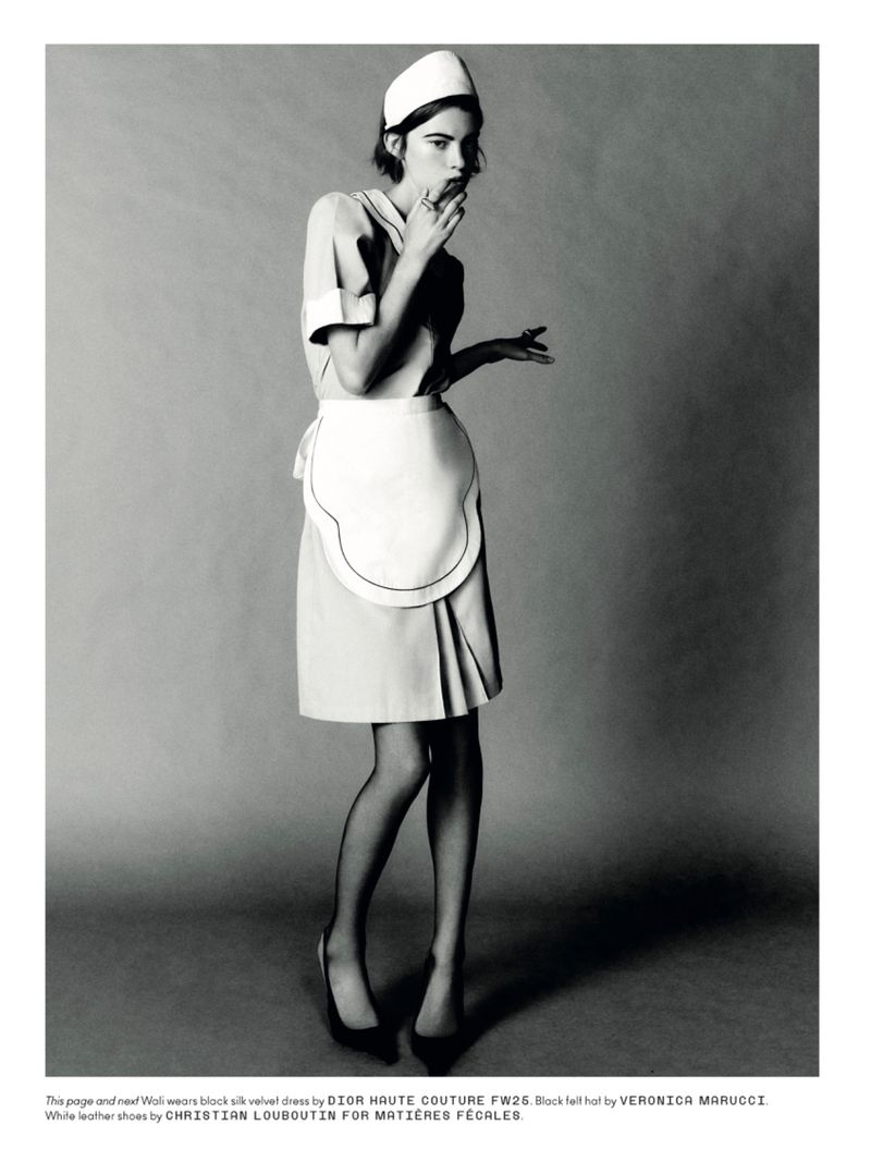 A young woman in a vintage nurse uniform poses in a studio, wearing black tights and high heels, set against a neutral gray background.