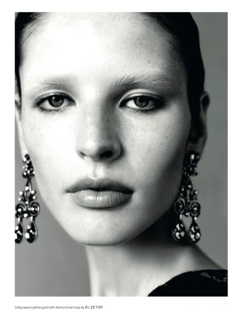 Close-up portrait of a woman wearing ornate dangle earrings and dark eyeliner against a grey background