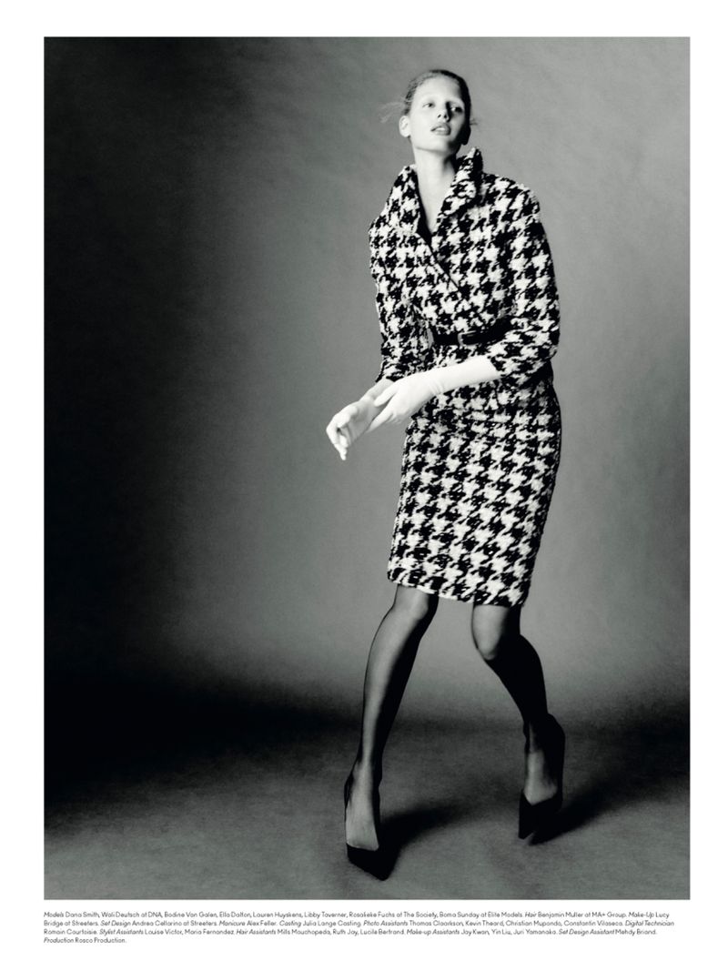 Fashion editorial shot of a model in a houndstooth skirt suit, black stockings and pumps, against a neutral backdrop in a photo studio.