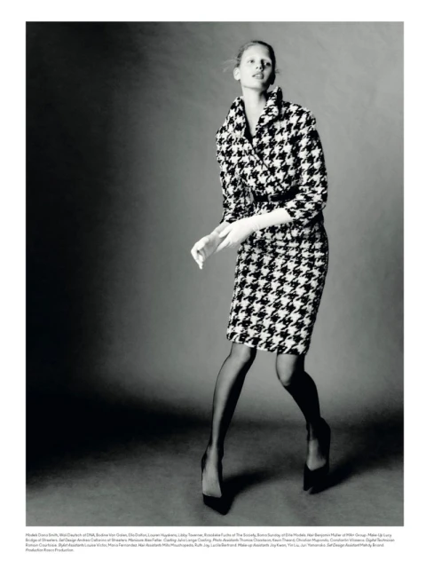 Fashion editorial shot of a model in a houndstooth skirt suit, black stockings and pumps, against a neutral backdrop in a photo studio.