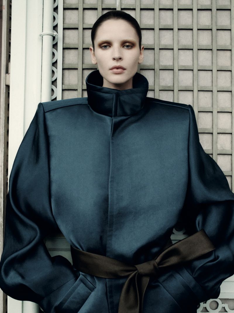 Woman in a blue satin jacket with a brown sash, posing against a neutral wall with symmetrical molding. Fashion editorial.