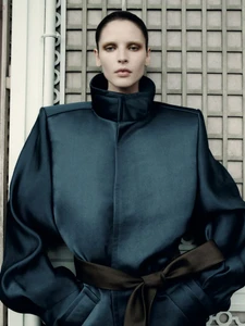 Model in a structured dark teal coat with a high collar and tied belt against a lattice wall.