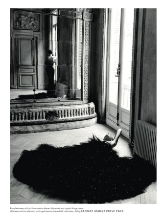 Model lying on wooden parquet floor in a voluminous black gown near a tall window and ornate mirror reflecting an interior.