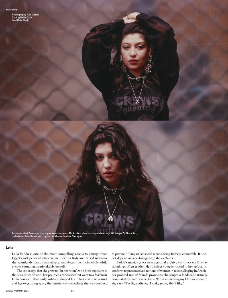 Portrait of a young woman with dark curly hair wearing a black top and silver jewelry against a textured backdrop in dim lighting.