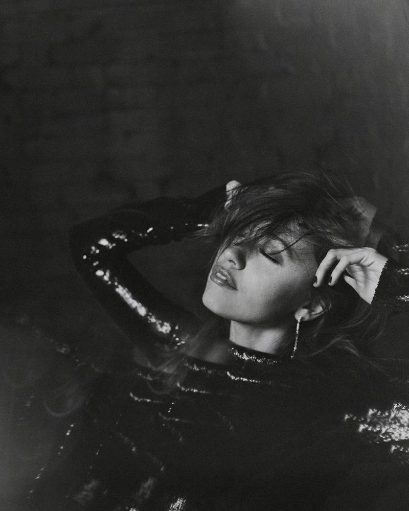 Adult female with eyes closed reclines wearing a dark sequined dress and dangling earrings in a monochrome studio portrait.