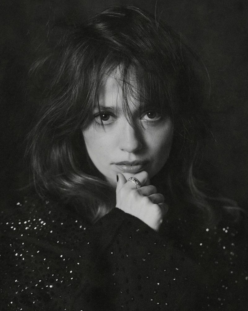 Portrait of a young woman with dark hair, wearing a black, sequined top, and holding her hand to her chin in a pensive pose.